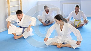 Team of athletes in kimono sits in a butterfly pose and practices stretching in sport gym