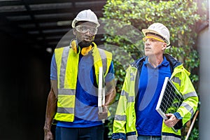 Team factory worker technician engineer men in green working suit dress and safety helmet talking and holding solar panel.