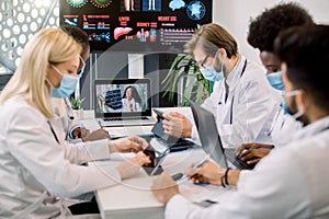 Team of five multiethnical doctors in protective masks, having video conference meeting in hospital with their young