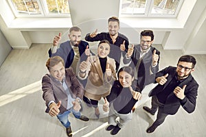 Team of happy business people standing in office, looking up, smiling and giving thumbs up