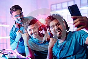 Team of happy cheerful professional gamers with headphones vlogging, live streaming, or taking selfie on mobile phone in front of