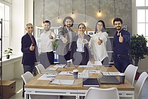 Team of happy employees giving thumbs up standing in office after corporate meeting