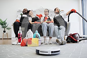 Team of housekeepers sitting on couch while robot vacuum cleaner cleans floor.