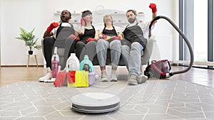 Team of housekeepers sitting on couch while robot vacuum cleaner cleans floor.