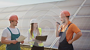 Team of industrial colleagues using tablet monitoring rows of photovoltaic solar panels at sunset. Business cooperation