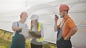 Team of industrial colleagues using tablet monitoring rows of photovoltaic solar panels at sunset. Business cooperation