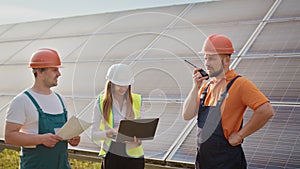 Team of industrial colleagues using tablet monitoring rows of photovoltaic solar panels at sunset. Business cooperation