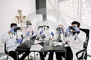 Team of multiethnic pharmacologists wearing protective white coats, gloves and vr goggles, working together on new