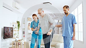 Team of nurses or social workers helping an old disabled man to walk with his crutches out of the nursing home room