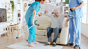 Team of nurses or social workers helping an old disabled man to walk with his crutches out of the nursing home room