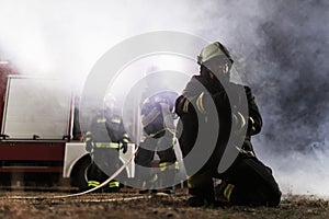 Team of professional firefighters holding water hose in front of a firetruck with smoke in the air. Half silhouette