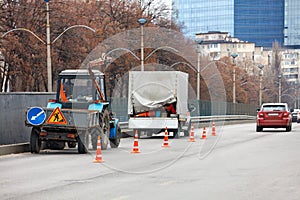 A team of road service workers on an old communal tractor and a truck are enclosing a section of the road with orange cones for