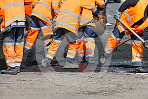 A team of road workers in orange reflective uniforms use shovels to scatter some of the asphalt to repair a section of the road on