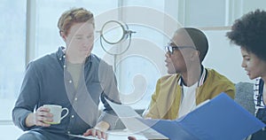 Team of three professionals discussing project plans in office lounge, with mugs and blue binder
