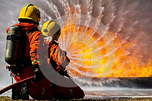 Team of two firefighters wearing full uniform with oxygen tank on back holding pipe spread water and face to wide huge orange hot