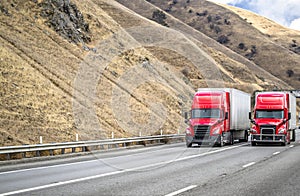 Team of two red big rigs semi trucks transporting cargo in dry van semi trailers running together on the multiline mountain