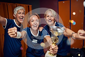 Team of two senior and one young women in dressing room, holding golden cup and feeling victorious