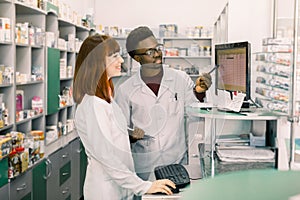 Team of two young apothecaries in pharmacy standing in front of shelves with drugs and working on computer making order