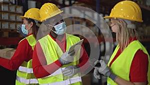 Team working together in warehouse doing inventory using digital tablet and loading delivery boxes while wearing face mask