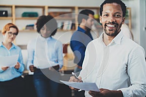 Teamwork concept in modern office.Young african businessman wearing white shirt holding papers at hands and standing