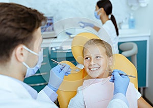 Male doctor in white coat with rubber gloves and protective mask treats teeth of smiling girl in chair