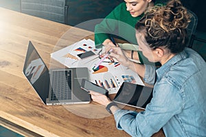 Teamwork. Two young business women sitting at table in front of laptop. On table is tablet computer and paper charts.