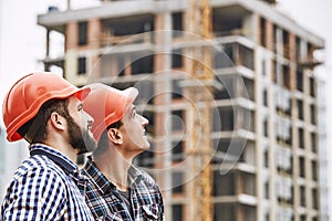 Teamwork. Two young and cheerful builders in red helmets are looking up and working at construction site