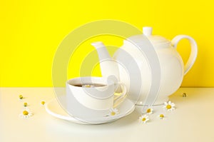 Teapot, cup of tea and chamomile on table against yellow background