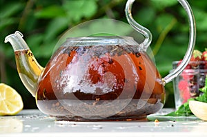 teapot full of tea on a background of green foliage on a wooden table, next to forest berries, raspberries and