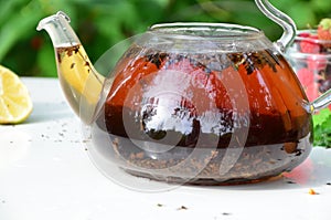 teapot full of tea on a background of green foliage on a wooden table, next to forest berries, raspberries and