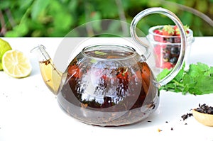 teapot full of tea on a background of green foliage on a wooden table, next to forest berries, raspberries and