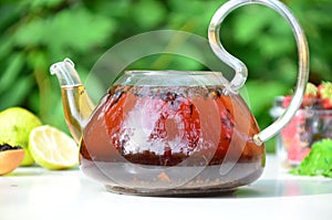 teapot full of tea on a background of green foliage on a wooden table, next to forest berries, raspberries and