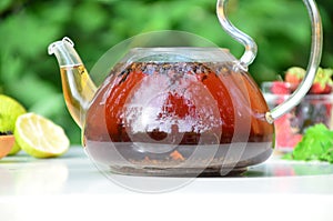 teapot full of tea on a background of green foliage on a wooden table, next to forest berries, raspberries and