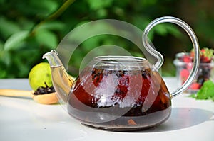 teapot full of tea on a background of green foliage on a wooden table, next to forest berries, raspberries and