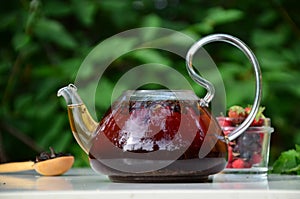 teapot full of tea on a background of green foliage on a wooden table, next to forest berries, raspberries and