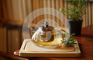 Teapot with herb tea on wooden cutting board with rosemary near, books on background, home cozy interior