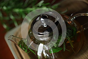 Teapot with herb tea on wooden cutting board with rosemary near, books on background, home cozy interior