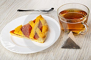 Spoon, piece of shortcrust pie with apricot jam in plate, cup with tea, tea bag on wooden table