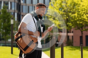 Tech gadgets for entertainment. Happy african american guy walking and using smartphone outdoors in ubran park area