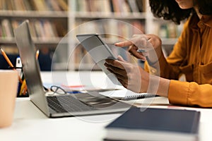 Tech and people concept. Woman using modern digital tablet while sitting in library at table with laptop computer
