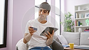 Tech-savvy young hispanic man relaxing on sofa, shopping online with credit card and touchpad indoors - portrait of serious but