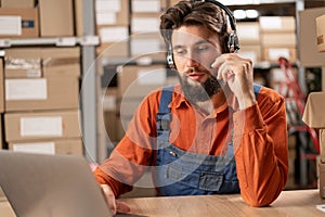 technical support operator at a delivery warehouse works at a laptop using a headset. Smiling handsome man talking to a