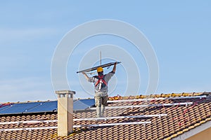 A technician lifts a solar panel over his head as he prepares to mount it on the roof of a red-tiled house