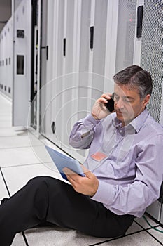 Mature technician sitting in data center beside cabinets holding tablet and smartphone, copy space