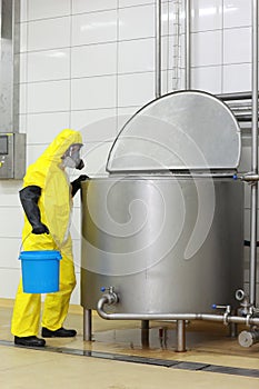 Technician in protective coveralls with blue bucket at opened industrial process tank