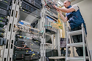 Technician standing on a ladder connects internet wires. The specialist works in the server room of the data center. Worker lays