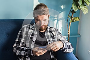 Technologies and leisure concept - Cheerful man sitting on the couch using his smartphone at home in the living room.