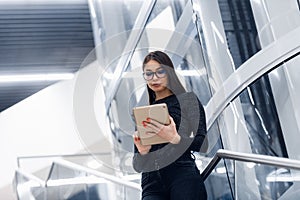 Technology, business and corporate concept - happy woman or student with tablet pc computer standing on stairs at modern