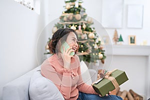 Technology. Communication. Christmas. Young woman is holding a gift box, talking on the mobile phone and smiling while sitting