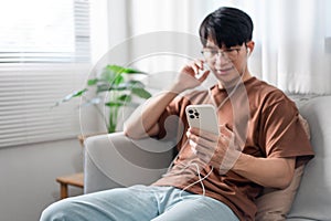 Technology Concept The person who wears glasses and headphones sitting on the grey couch and listening music on his smartphone